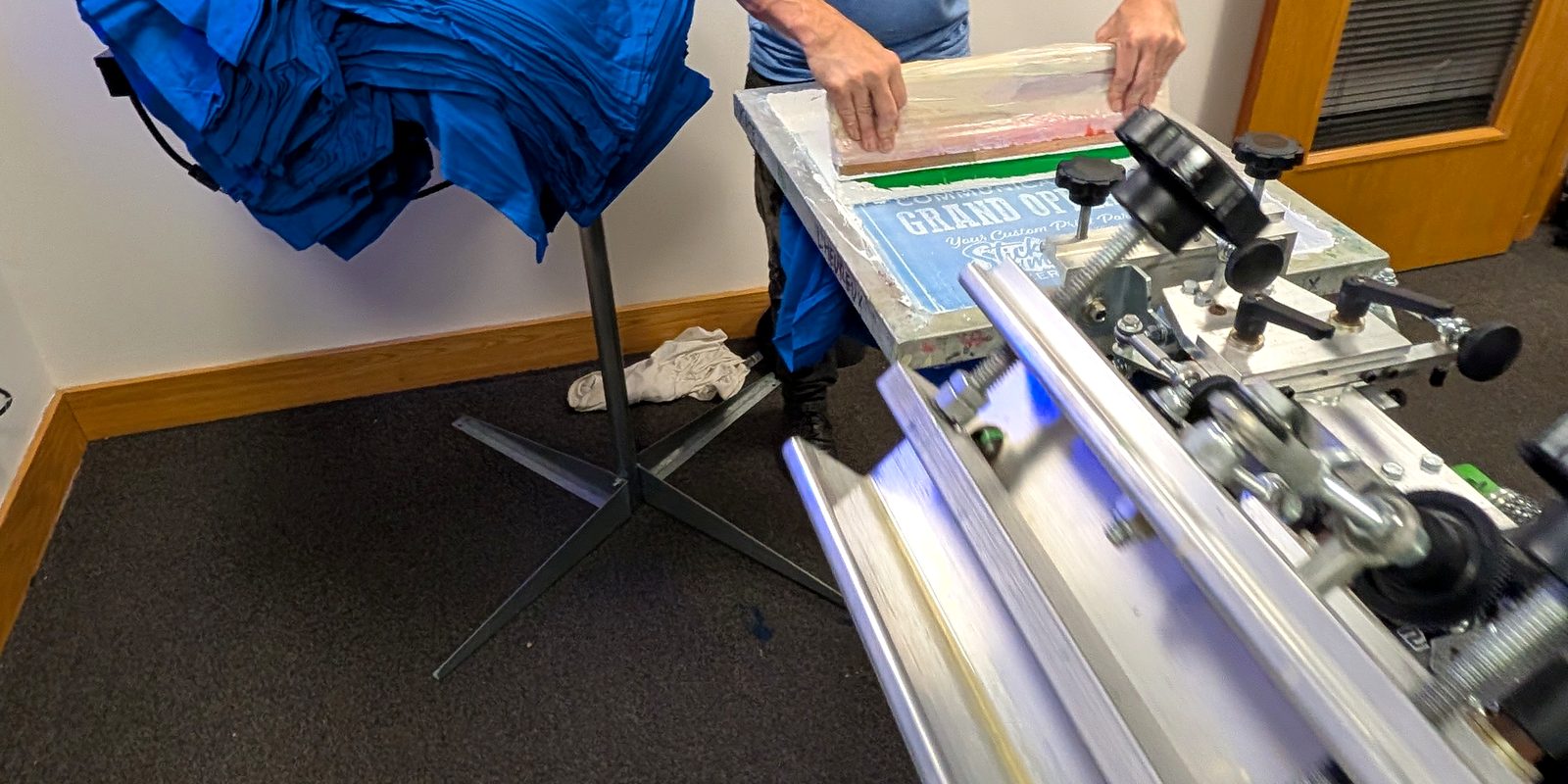 Pulling a squeegee across a screen on a manual press, with a stack of blue shirts ready to print